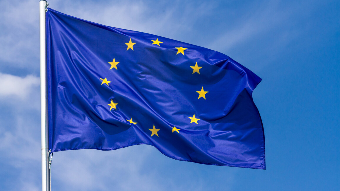 An EU flag is blowing in the wind. The sky in the background is blue and only slightly cloudy.