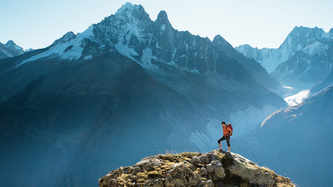 Ein Mann steht auf einer Bergspitze. Im Hintergrund ist ein Gebirgspanorama zu sehen.