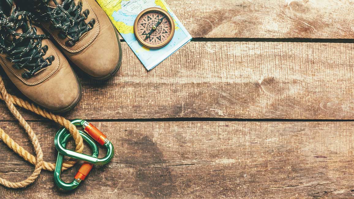 Hiking shoes, a map, a compass and a rope with snap hooks are lying on the wooden floor.