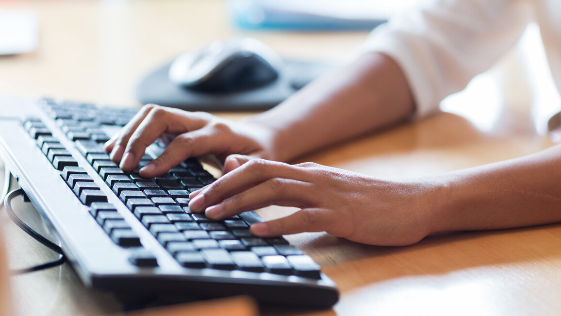Zwei Hände tippen auf einer schwarzen Computer-Tastatur, die auf einem Holztisch steht. 